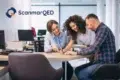 A team of three people collaborates around a desk in a bright office. Two women and a man are focused on documents, smiling. ScanmarQED logo in the corner.