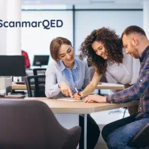 A team of three people collaborates around a desk in a bright office. Two women and a man are focused on documents, smiling. ScanmarQED logo in the corner.