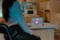 A woman in a white blouse sits at a kitchen counter using a tablet displaying smart home controls. The modern kitchen background is softly lit.