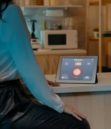 A woman in a white blouse sits at a kitchen counter using a tablet displaying smart home controls. The modern kitchen background is softly lit.