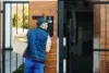 A delivery person in a blue uniform and cap holds a package while speaking into an intercom at a modern gate, conveying a sense of purpose and focus.