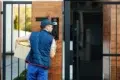 A delivery person in a blue uniform and cap holds a package while speaking into an intercom at a modern gate, conveying a sense of purpose and focus.