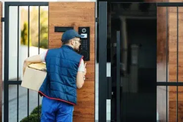 A delivery person in a blue uniform and cap holds a package while speaking into an intercom at a modern gate, conveying a sense of purpose and focus.