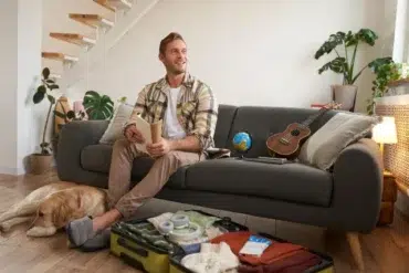 A man sits on a grey sofa with a travel book, looking happy. An open suitcase, globe, and ukulele are nearby. A dog rests on the floor. The room is bright and cozy, with houseplants and a small staircase in the background.