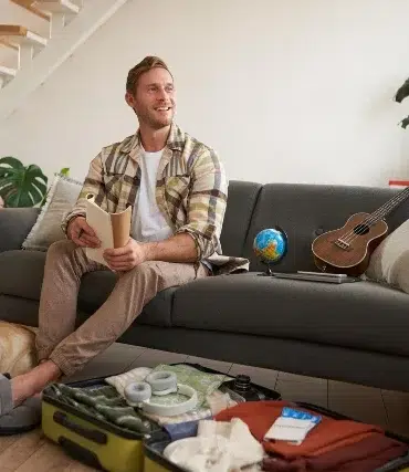 A man sits on a grey sofa with a travel book, looking happy. An open suitcase, globe, and ukulele are nearby. A dog rests on the floor. The room is bright and cozy, with houseplants and a small staircase in the background.