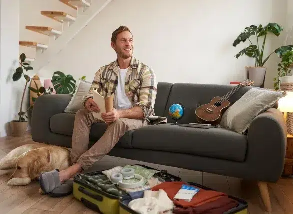 A man sits on a grey sofa with a travel book, looking happy. An open suitcase, globe, and ukulele are nearby. A dog rests on the floor. The room is bright and cozy, with houseplants and a small staircase in the background.