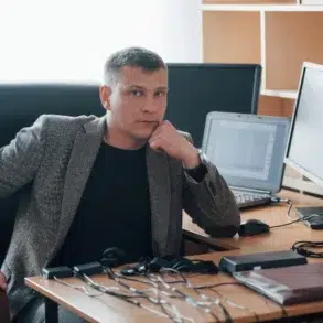 A man in a gray blazer sits in an office at a wooden desk with multiple monitors, a laptop, and electronic gadgets, appearing contemplative.