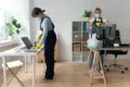 Two people are cleaning an office, wearing masks and gloves for safety. They are tidying desks with cleaning supplies, conveying a diligent, professional tone.