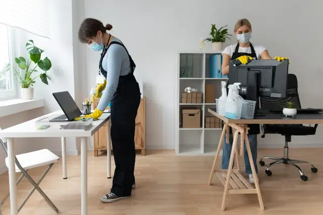 Two people are cleaning an office, wearing masks and gloves for safety. They are tidying desks with cleaning supplies, conveying a diligent, professional tone.