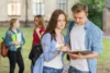 Two students discuss a book outdoors while other students walk by in a university setting. Bright and lively atmosphere.