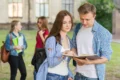 Two students discuss a book outdoors while other students walk by in a university setting. Bright and lively atmosphere.