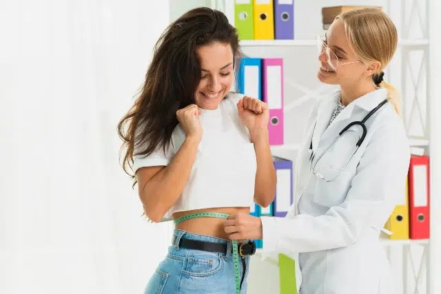 A woman smiles as a doctor measures her waist with a tape measure. They're standing in a bright room with colorful folders, conveying a positive, supportive atmosphere.