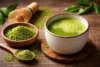 “What Does Matcha Taste Like” showing a frothy cup of bright green matcha tea with matcha powder, a bamboo whisk, and fresh tea leaves on a wooden table.