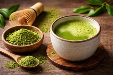 “What Does Matcha Taste Like” showing a frothy cup of bright green matcha tea with matcha powder, a bamboo whisk, and fresh tea leaves on a wooden table.