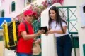 A delivery person in a red uniform holds a package while a woman signs on a tablet outside a house with colorful walls and flowers, conveying a friendly transaction.