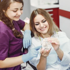A dentist and patient are smiling and examining a dental model. The dentist, in purple scrubs, points at the model while the patient holds it.