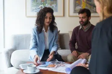 Three people engaged in a discussion around a coffee table with documents. A woman in a blue blazer gestures towards images; a calm, focused atmosphere.