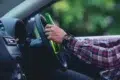 A person in a plaid shirt holds a green bottle while steering a car, suggesting the risky behavior of drinking and driving. The mood is tense.