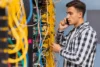 A man in a plaid shirt talks on his phone while examining a server rack filled with tangled yellow cables, conveying a sense of focus and urgency.