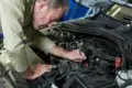 A mechanic works on a car engine, using a wrench to adjust components under the hood, surrounded by various engine parts.
