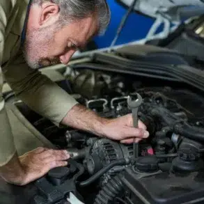 A mechanic works on a car engine, using a wrench to adjust components under the hood, surrounded by various engine parts.