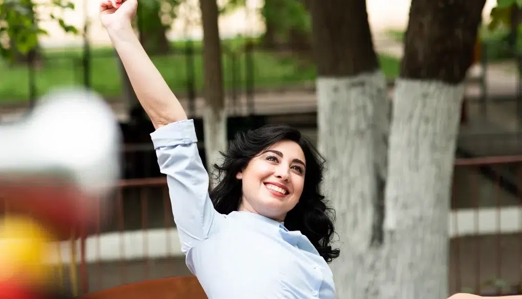 A person raises their arm in celebration while sitting outdoors, surrounded by trees and a blurred playground in the background.