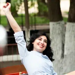 A person raises their arm in celebration while sitting outdoors, surrounded by trees and a blurred playground in the background.