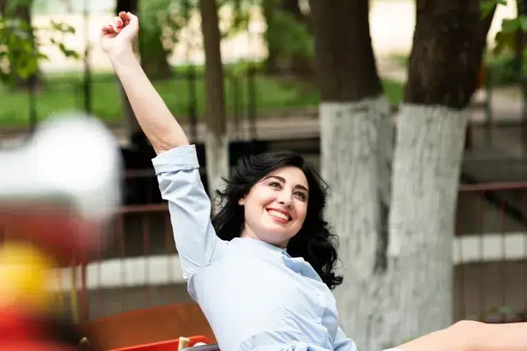 A person raises their arm in celebration while sitting outdoors, surrounded by trees and a blurred playground in the background.