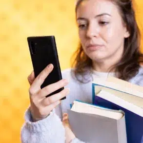 A woman holding several books looks at her smartphone, appearing focused. She stands against a bright yellow background, wearing a soft blue sweater.