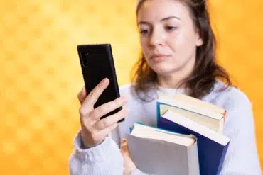 A woman holding several books looks at her smartphone, appearing focused. She stands against a bright yellow background, wearing a soft blue sweater.