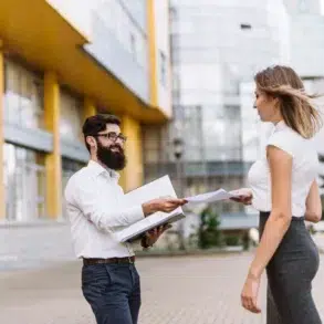 A bearded man and a woman exchange documents outdoors, smiling. They stand in front of a modern glass building. The scene feels professional and friendly.