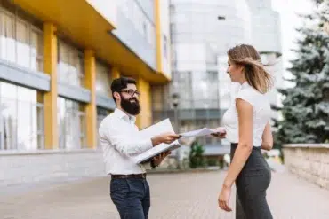 A bearded man and a woman exchange documents outdoors, smiling. They stand in front of a modern glass building. The scene feels professional and friendly.
