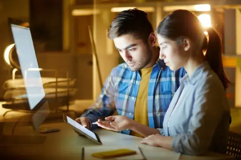 Two individuals are engaged in discussion at a desk, with a laptop and tablet in a softly lit workspace.
