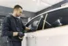 A person in a dark outfit applies masking tape along a car window in a well-lit garage, preparing for painting or detailing.