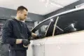 A person in a dark outfit applies masking tape along a car window in a well-lit garage, preparing for painting or detailing.