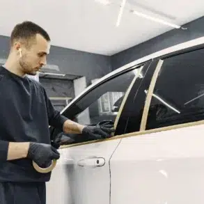 A person in a dark outfit applies masking tape along a car window in a well-lit garage, preparing for painting or detailing.