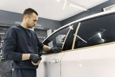 A person in a dark outfit applies masking tape along a car window in a well-lit garage, preparing for painting or detailing.