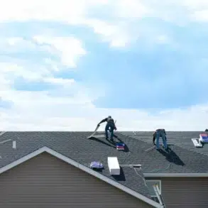 Four workers are installing roofing shingles on a house under a bright blue sky. The scene conveys industriousness and teamwork.