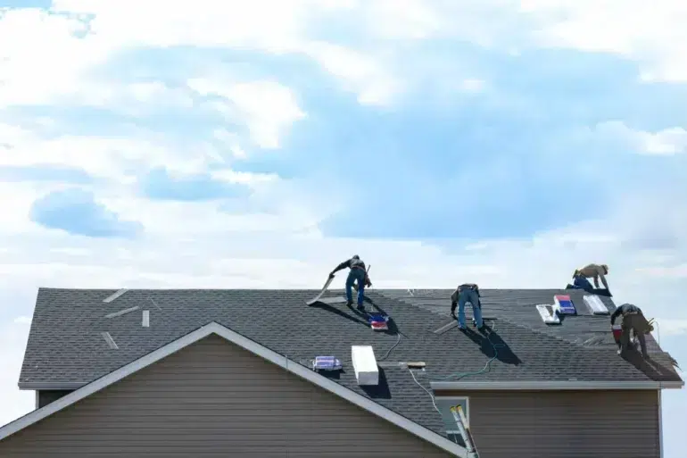 Four workers are installing roofing shingles on a house under a bright blue sky. The scene conveys industriousness and teamwork.