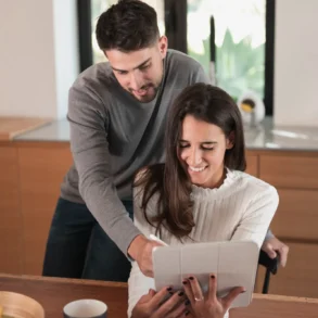 A smiling woman in a cozy kitchen holds a tablet while a man leans over, pointing at the screen. They appear engaged and happy, sharing a moment.