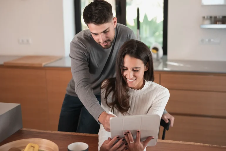 A smiling woman in a cozy kitchen holds a tablet while a man leans over, pointing at the screen. They appear engaged and happy, sharing a moment.