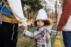 Child dressed in a plaid coat and white hat holding hands with two adults outdoors in a park during autumn.