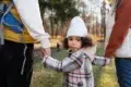 Child dressed in a plaid coat and white hat holding hands with two adults outdoors in a park during autumn.