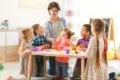 A teacher engages a diverse group of children at a table, playing with colorful playdough in a bright classroom setting.