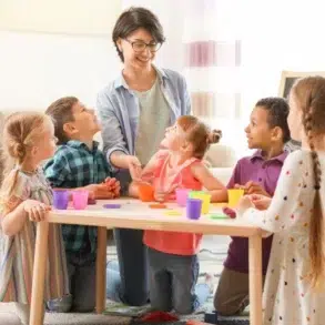 A teacher engages a diverse group of children at a table, playing with colorful playdough in a bright classroom setting.