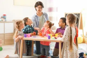 A teacher engages a diverse group of children at a table, playing with colorful playdough in a bright classroom setting.