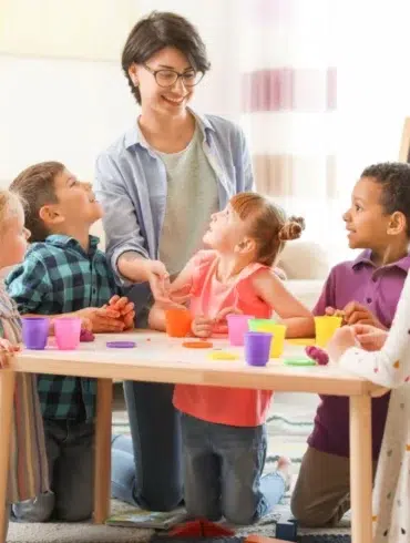 A teacher engages a diverse group of children at a table, playing with colorful playdough in a bright classroom setting.