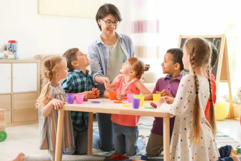 A teacher engages a diverse group of children at a table, playing with colorful playdough in a bright classroom setting.