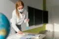 A masked woman cleans a classroom desk with a spray bottle and cloth, conveying hygiene and safety. A globe and chalkboards are visible in the background.