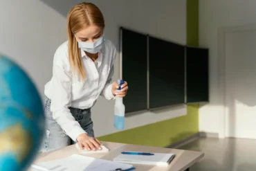 A masked woman cleans a classroom desk with a spray bottle and cloth, conveying hygiene and safety. A globe and chalkboards are visible in the background.